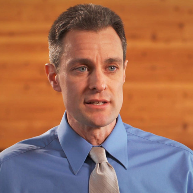 Damian Costello sits in front of a mid-tone wood background, wearing a blue collared shirt and silver tie. He has sandy brown hair and blue eyes and looks to be speaking to someone off-camera.