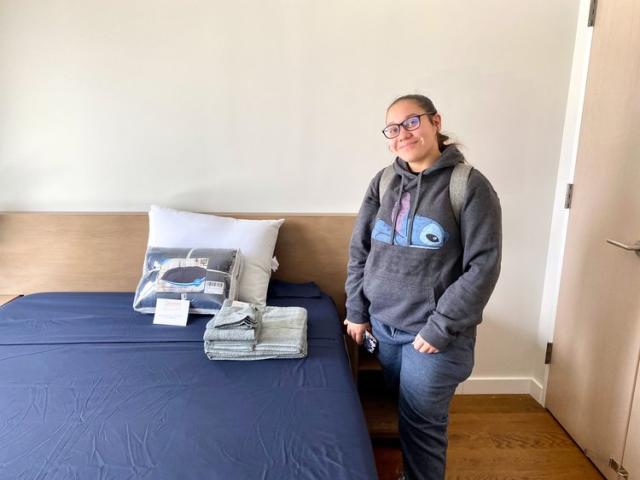 CUNY student Odalys P. wears a grey hoodie, glasses, and jeans while smiling next to a bed with extra linens and towels on it.
