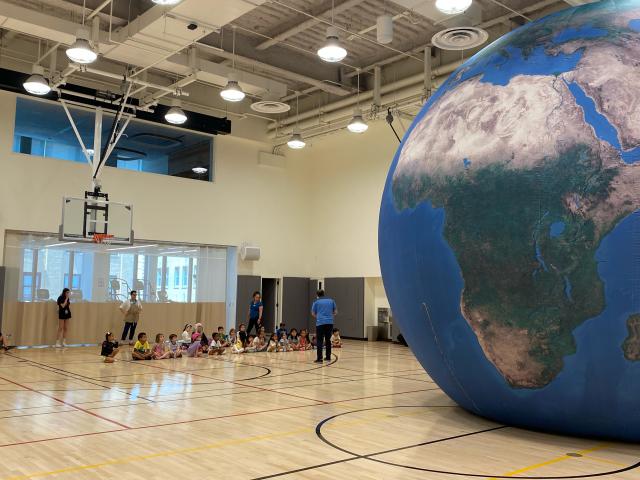A group of children sitting in the Trinity Commons gymnasium sit before a large inflatable Earth globe learning a science lesson.