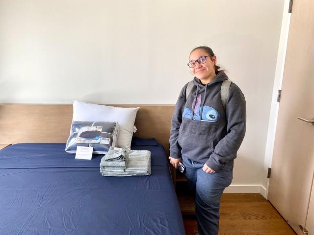 CUNY student Odalys stands in front of her dorm room bed and bedding after moving into her new home at the NCS Scholars program.