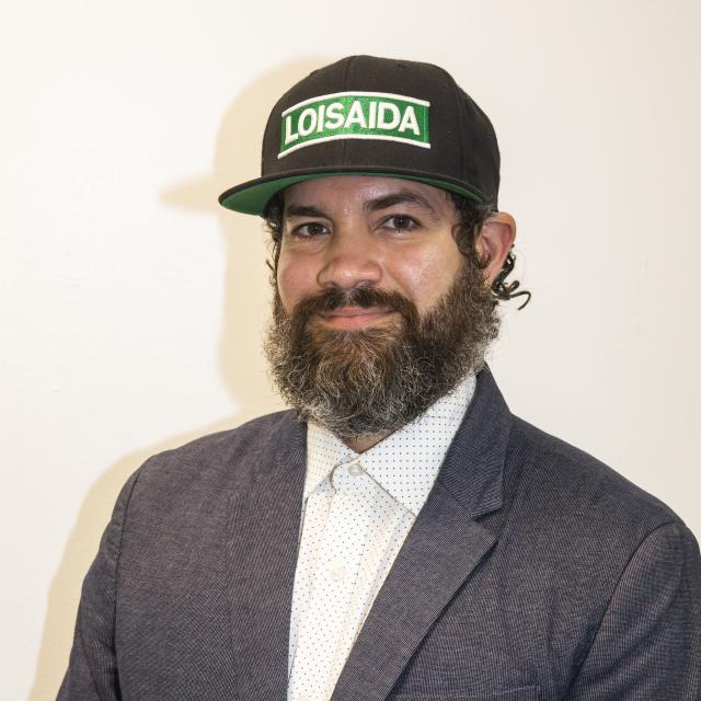 Alejandro Epifanio wears a hat that reads "Loisaida" with a grey blazer and collared shirt.