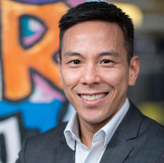 Kelsey Louie smiles with a suit and open collar in front of graffiti.