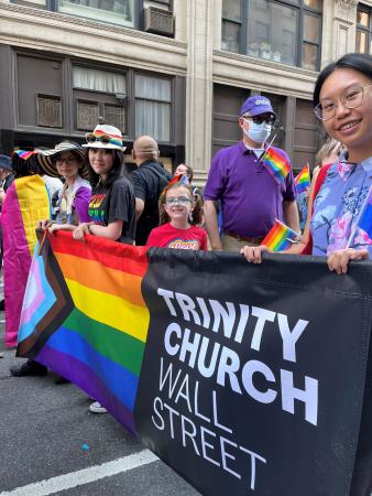Three young women and a girl hold a banner with the Progress Pride Flag and the Trinity Church Wall Street logo as they walk in the 2022 Pride march in New York City.