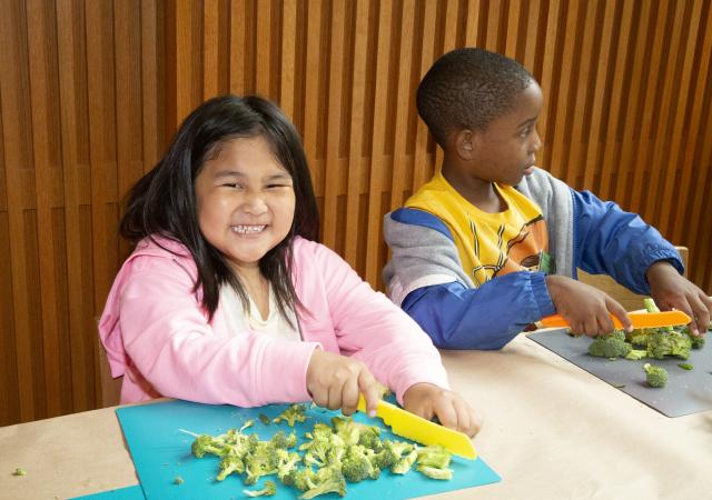 A girl smiles at the camera while chopping broccoli during the 2023 Children's Summer Program