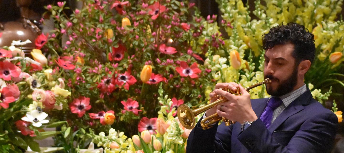 Man plays piccolo trumpet in front of a burst of spring flowers