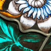 A close-up photo of stained glass depicting a white and blue flower with vivid green leaves