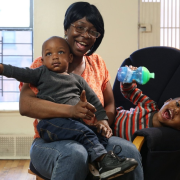 A Black mother wearing glasses and her hair in a black bob smiles with her two children. One child smiles and leans over with his sippy cup in hand, while the other sits in her lap with a serious expression and points to the left.