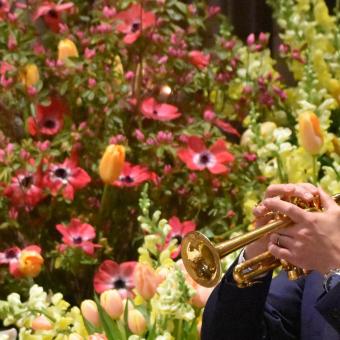 Man plays piccolo trumpet in front of a burst of spring flowers