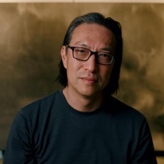 Makoto Fujimura stands in front of an ochre background, wearing glasses and a black crewneck shirt. He smiles slightly with his head tilted to the right.