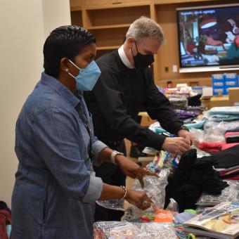 A brown skinned woman with short hair wearing a blue mask & white man wearing priest's garb and a mask pack books, clothing, and other supplies. The room is full of boxes and boxed food items.