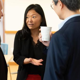 An Asian woman is in conversation with a White man and Asian Man. She has dark hair and is wearing a black cardigan, black lace-lined blouse, and red skirt. The White man has white hair and is wearing a dark grey suit. The Asian man wears glasses, a navy suit, and is holding a white coffee cup.