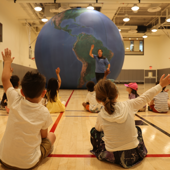 Children in the Trinity Commons gym raise their hands in front of an inflatable globe as part of a science lesson during the 2023 Children's Summer Program