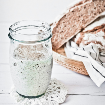 An image of sourdough starter in a glass jar, with a sliced loaf of bread in the background.