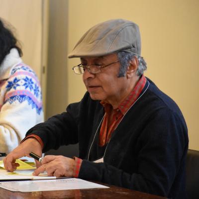 A man wearing a beige ivy cap sits at table with paperwork in front of him. He is facing left, looking down.