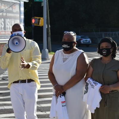 A masked man is speaking into a megaphone. Two masked women stand to his left.