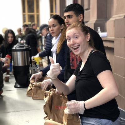 Volunteers stand in line serving Brown Bag Lunch to guests. A female volunteer looks directly into camera and gives a thumbs up.