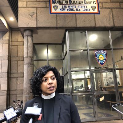 The Rev. Winnie Varghese stands with two microphones pointed at her.