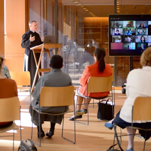 Members of the congregation sit in chairs with WebEx screen and Vicar speaking at Discovery