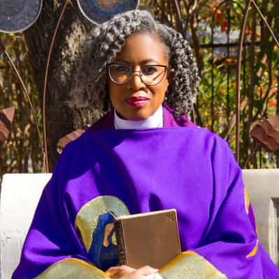 The Rev. Dr. Wilda C. Gafney, wearing a purple robe, sits in the sun, holding a Bible