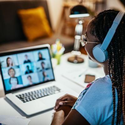 A young woman with braids is on a video call, wearing glasses and headphones. Her back is to us, while the computer screen is blurred.
