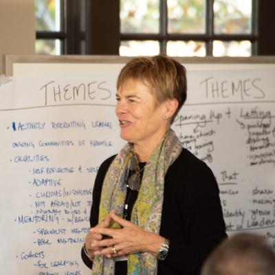 A white woman wearing a green scarf speaks to a group. Behind her is writing on a white poster on themes for leadership development.