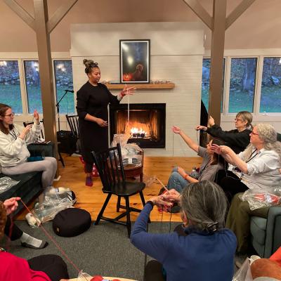 Parishioners on a Women's Retreat learn to knit. Four women are shown, sitting on couches in front of an instructor and a roaring fire at Trinity Retreat Center.