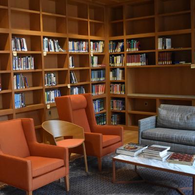 View of a corner of the Lina Lowery Library in Trinity Commons. There are wooden built-in bookshelves, two orange club chairs, a low gray couch and a marble-topped coffee table on a patterned gray area rug. 