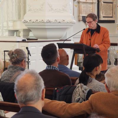Man in an orange jacket with short dark hair and glasses reads poetry from a lectern in front of an audience at St. Paul's Chapel.