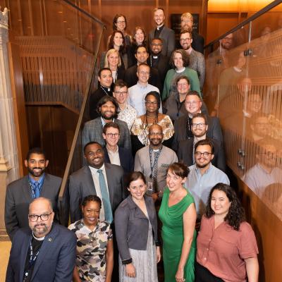 The 2022 Trinity Leadership Fellows cohort stands on a staircase at the Trinity Commons, smiling up at the camera. The multiracial group of men, women, and nonbinary people are dressed smartly, with some in suits, dresses, or clerical garb.