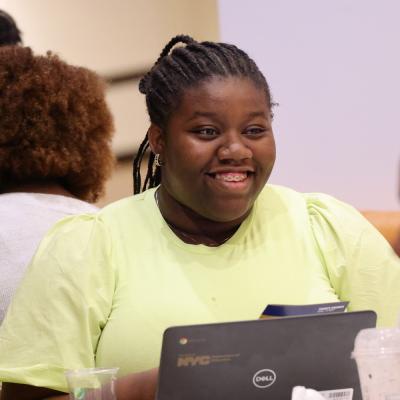 A young Black woman with braces smiles. She wears her hair in cornrows and dons a lime-colored shirt. She sits in front of a Dell laptop with a "Property of NYC Department of Education" logo on it.