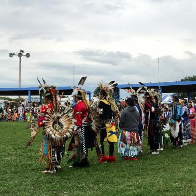 Sioux pow-wow: many adults wearing traditional, brightly colored garments including feather headwear and holding drums stand in a line on a grassy field.