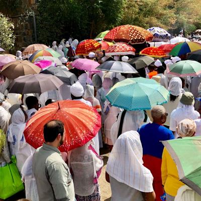 A crowd of people stand on a path. Many hold colorful umbrellas or wear scarves to protect themselves from the sun.