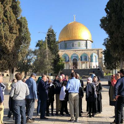 A group of people stand in a plaza under a bright blue, cloudless sky on a sunny day. They are visiting an historic temple in the Holy Land with a golden dome.
