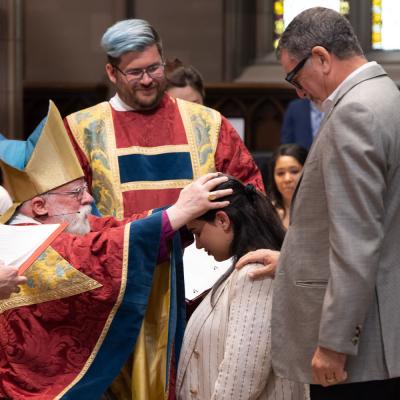 Bishop Dietsche lays hands on confirmand's brunette hair as she kneels to receive a blessing. Father Matt and a man with his hand on her shoulder fill out the group.