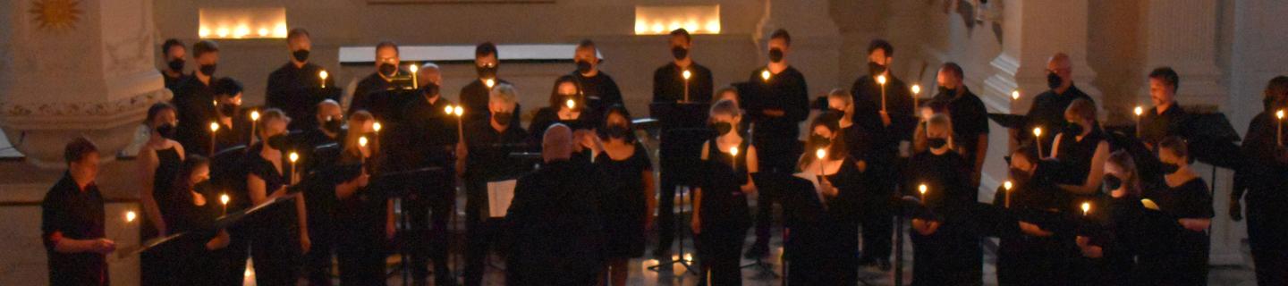 Men and women of The Choir of Trinity Wall Street standing in St. Paul's Chapel, wearing all black, holding candles and singing. 